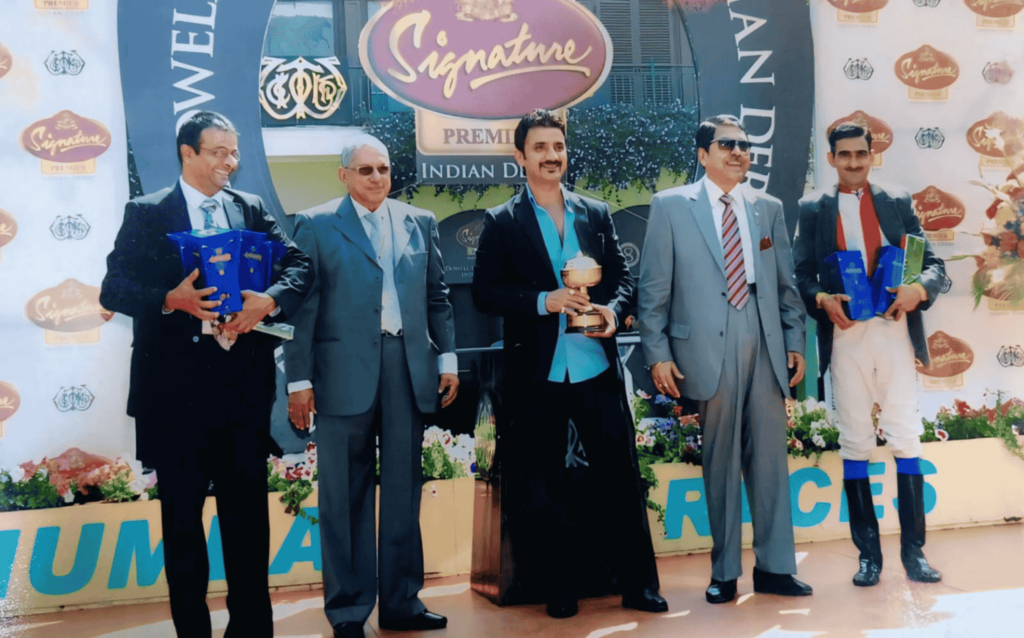 Sudheer Chauhan holding a trophy at the Signature Indian Derby award event, surrounded by race officials and jockeys in formal attire.